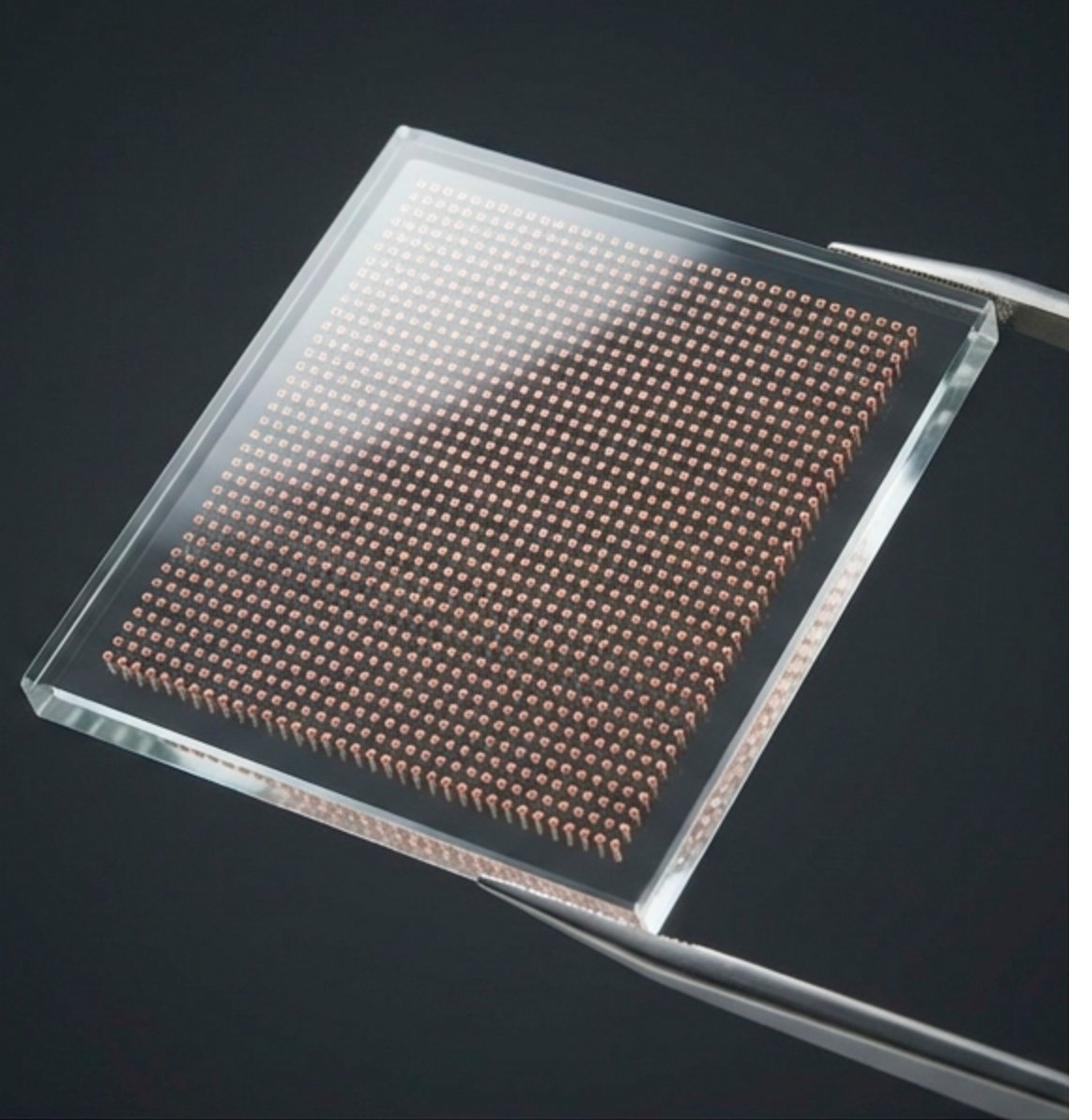 TGV glass interposer - through-glass vias filled with copper showing dense via array in transparent glass substrate held by tweezers